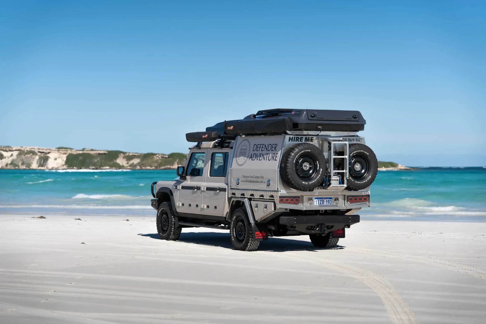 Defender Adventure 4WD Camper am Strand mit Dachzelten und Ersatzreifen vor türkisblauem Meer