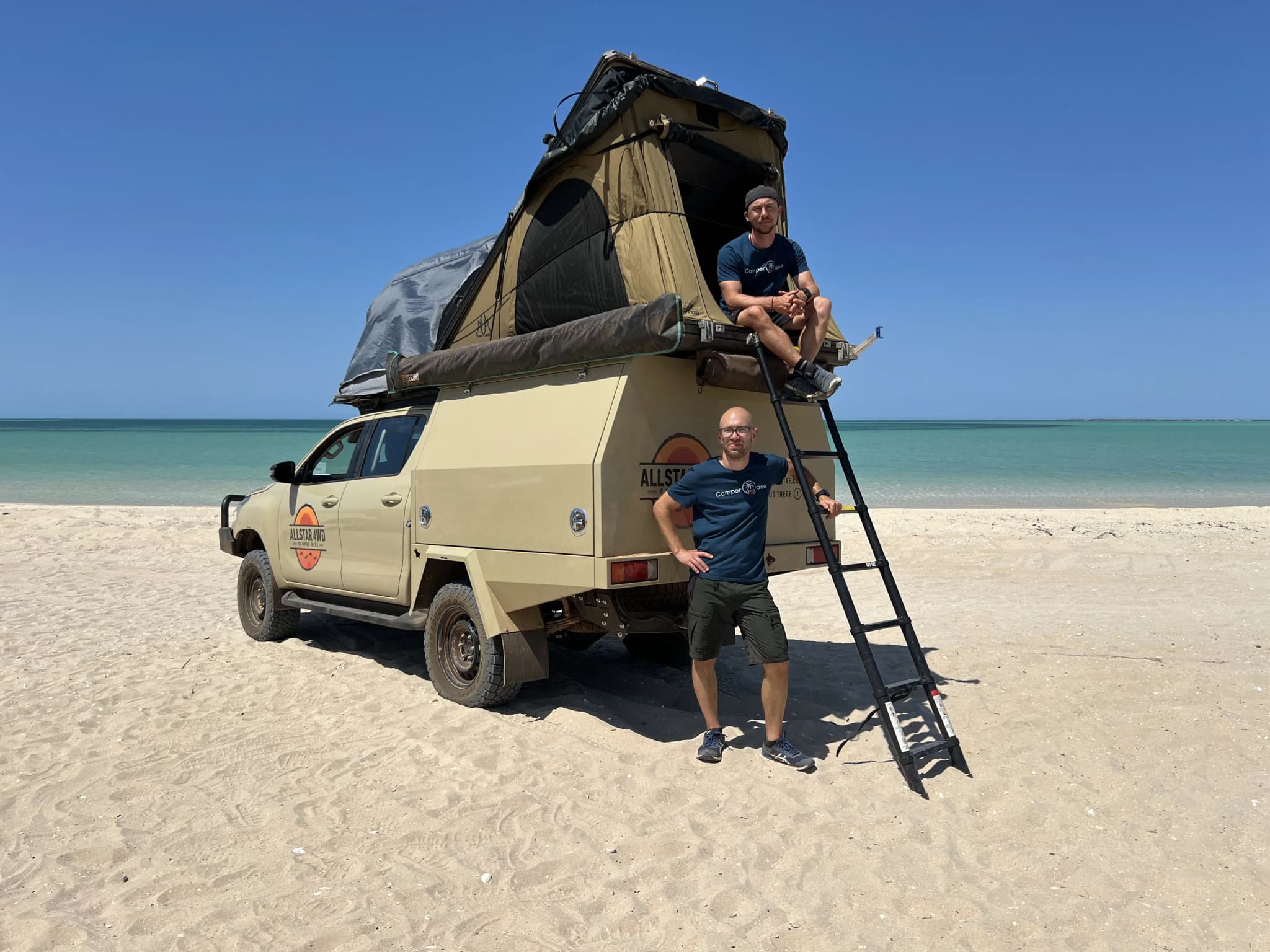 zwei Herren mit einem Allradcamper am Strand, einer Sitzend im Dachzelt, der andere neben der Leiter stehend