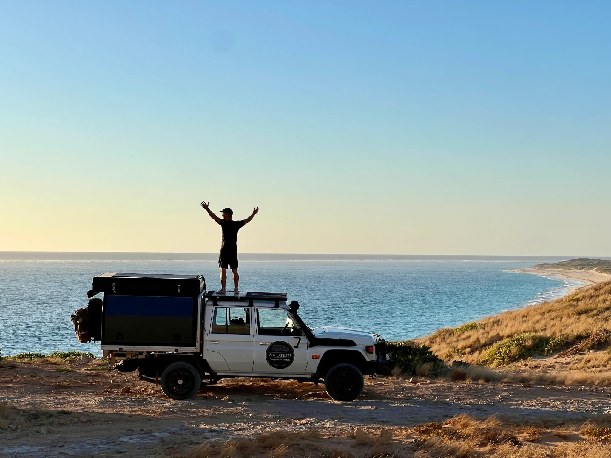 Person steht auf Allradcamper mit Blick auf Küste im Sonnenuntergang in Westaustralien und reißt die Arme nach oben