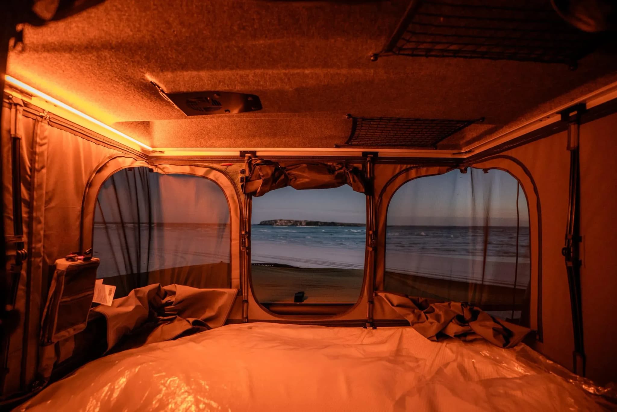 Innenansicht des geöffneten Dachzelts im Defender Adventure Ineos Quartermaster mit Bett, warmem Licht und Blick auf den Strand
